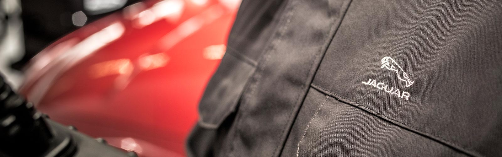 Close-up of embroidered Jaguar logo on a black jacket, with a blurred red car in the background.