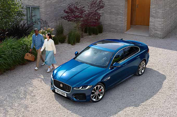 A blue luxury sedan parked on a gravel driveway with two people walking nearby, set against a modern brick building.