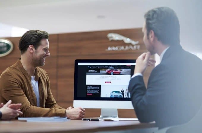 Two men discussing in front of a computer displaying a car dealership website with Jaguar branding.
