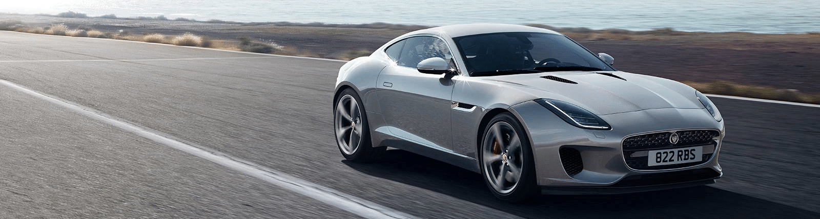 Silver sports car driving on an open road near the coast, showcasing sleek design and speed.