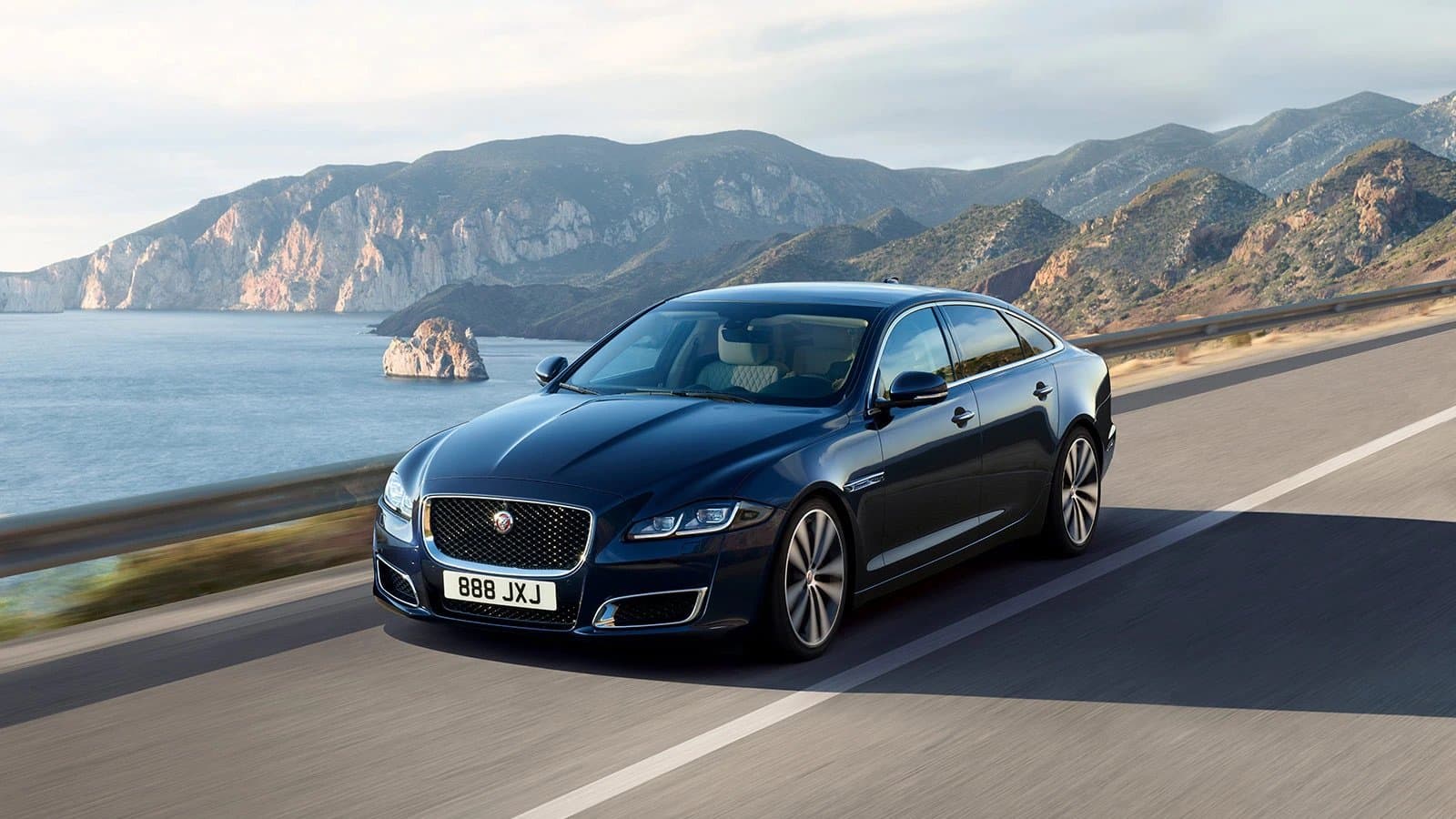 Sleek blue luxury sedan driving along a coastal highway with scenic mountains and ocean views.