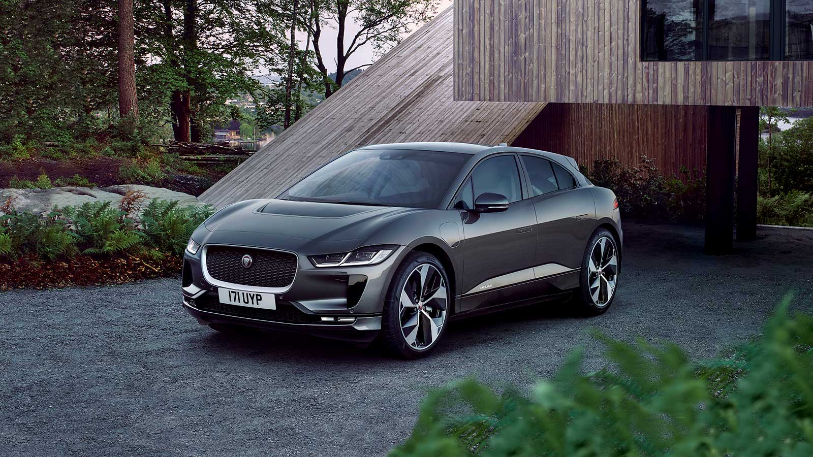 Sleek gray Jaguar I-PACE parked by a modern wooden house surrounded by trees.