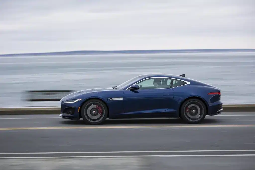 Dark blue sports car driving along a coastal road, ocean visible in the background.
