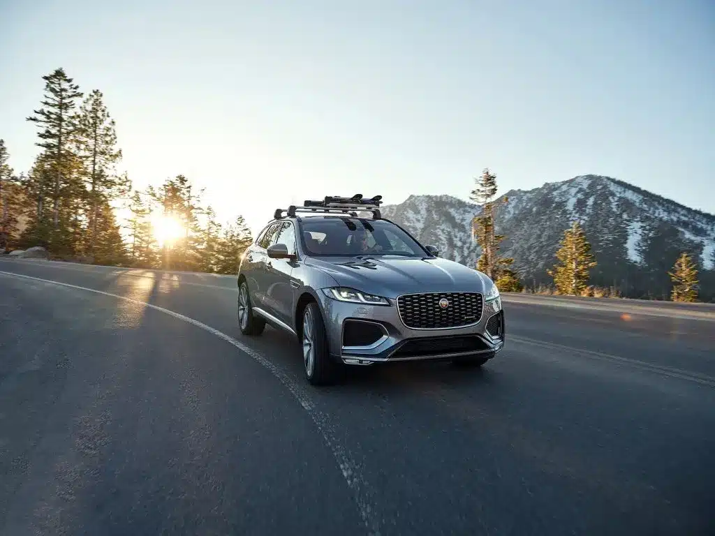 SUV driving on a curved mountain road at sunrise, with snow-capped peaks and evergreen trees.