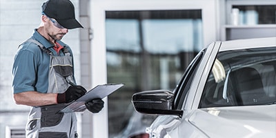 Mechanic in uniform checks a list on a clipboard beside a white car in a garage setting.