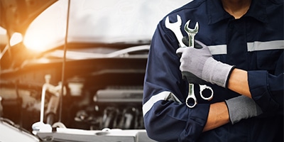 Mechanic holding wrenches in front of an open car hood, wearing gloves and a uniform.